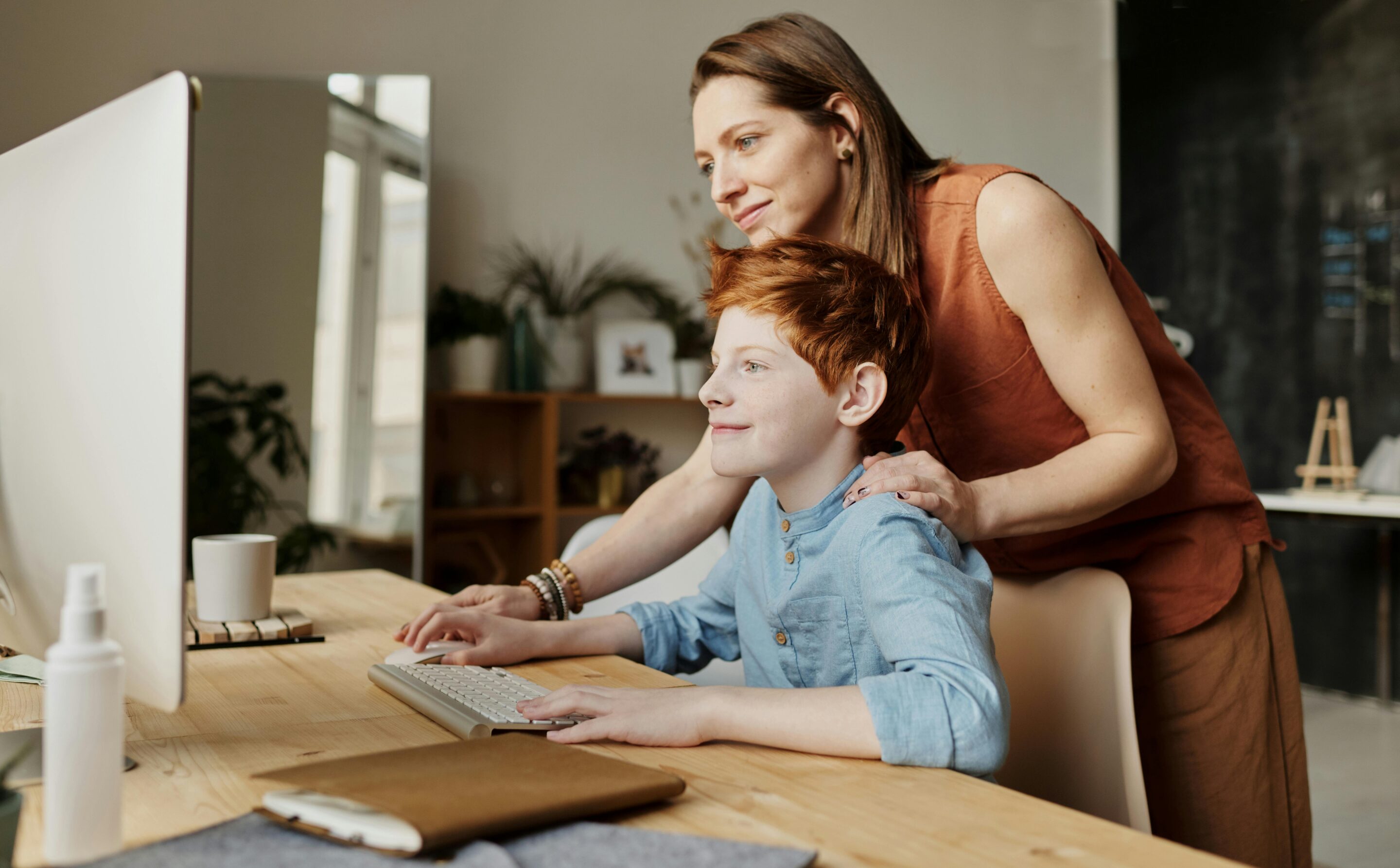 redhead child sitting at a desk on a computer with a woman with her hand on his shoulder