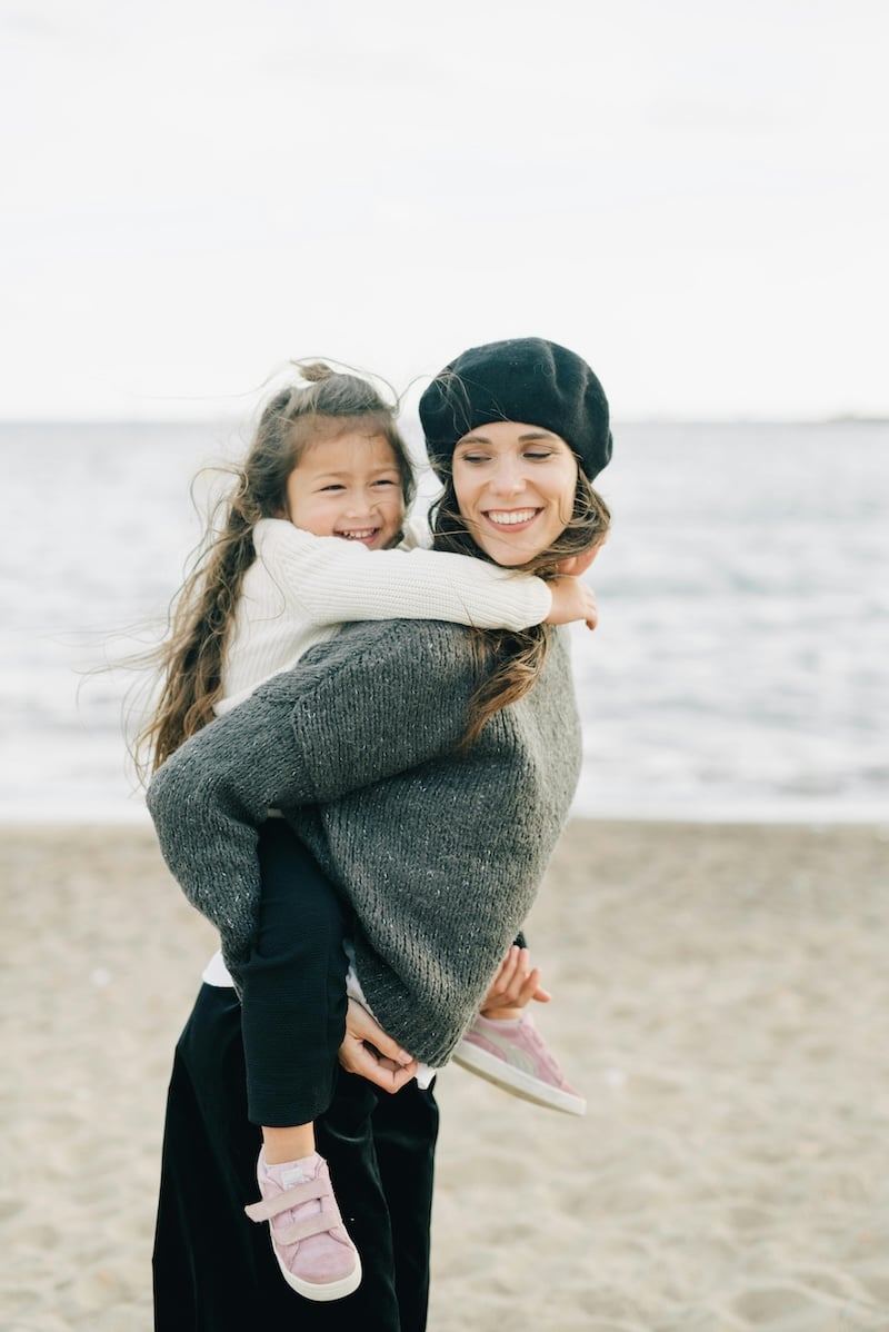 Women in grey sweater with long dark hair wearing a beret giving a brunette child with long hair a piggy back ride